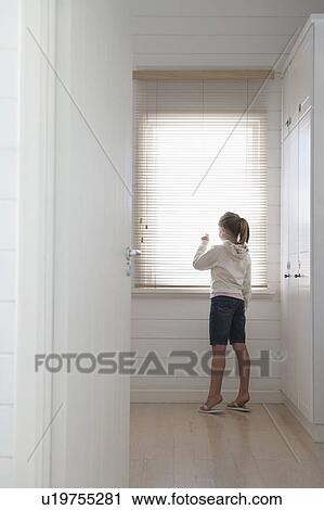 Stock Image - Young girl peering through blinds standing at window, full length. Fotosearch