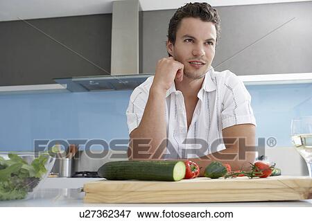 Stock Photo - Young man leaning on kitchen counter near cutting board with vegetables, hand on chin. Fotosearch