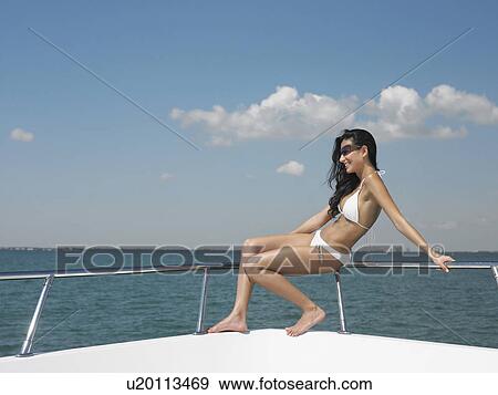 Stock Photo - Young woman in bikini sitting on bow of yacht, side view. Fotosearch