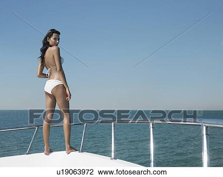 Stock Image - Young woman in bikini standing on bow of yacht, looking at camera, back view. Fotosearch