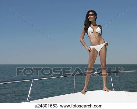 Stock Image - Young woman in bikini standing on bow of yacht, portrait. Fotosearch