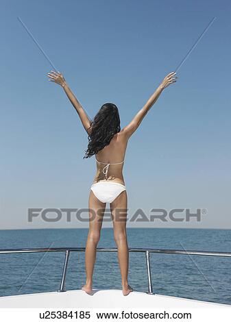 Stock Photography - Young woman in bikini standing on bow of yacht, outstretching arms, back view. Fotosearch