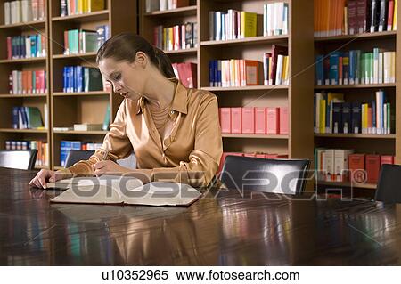 Stock Photography - Young woman reading book at desk in library. Fotosearch