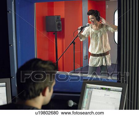 Stock Image - Young woman singing in studio, technician in foreground. Fotosearch