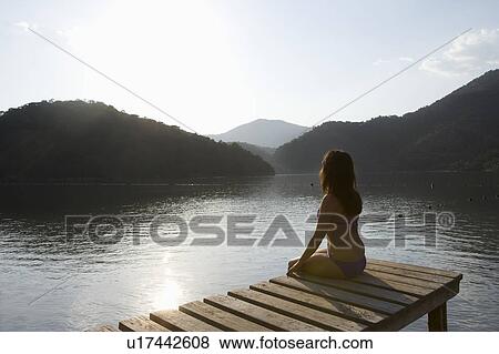 Stock Photo - Young woman sits on lakeside jetty at dawn. Fotosearch