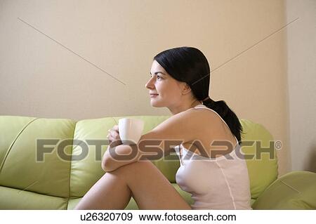 Stock Photo - Young woman sits on sofa, side view holding cup. Fotosearch
