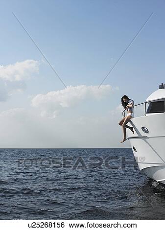 Young woman sitting at edge of yacht with legs dangling overboard, side view View Large Photo Image Stock Photograph - Young woman sitting at edge of yacht with legs dangling overboard, side view. Fotosearch