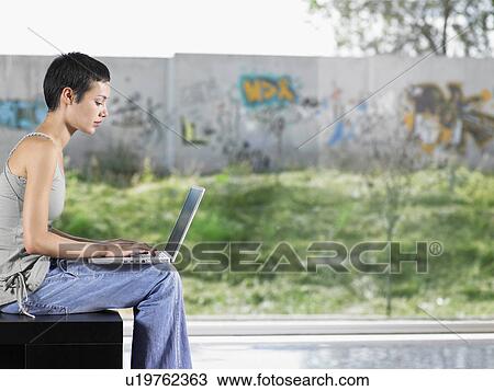 Stock Image - Young woman sitting down using laptop indoors. Fotosearch