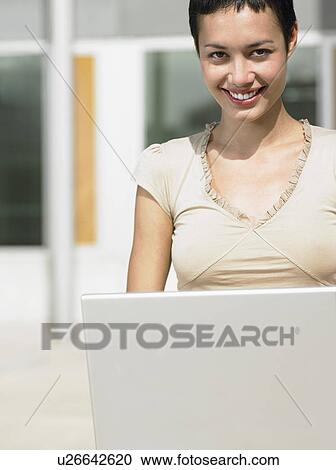 Stock Image - Young woman sitting in plaza courtyard using laptop, portrait. Fotosearch
