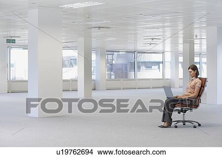 Picture - Young woman sitting in swivel chair, using laptop, in empty office space, side view. Fotosearch