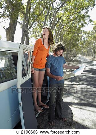 Stock Image - Young woman standing in camper van door, man looking at map, at side of road. Fotosearch