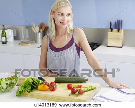 Stock Photo - Young woman standing in kitchen, hands on counter near cutting board with fresh produce. Fotosearch