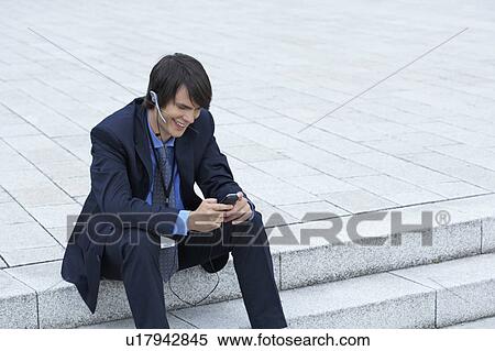 Stock Photography - Businessman sitting on steps with headset looking at his mobile laughing.. Fotosearch