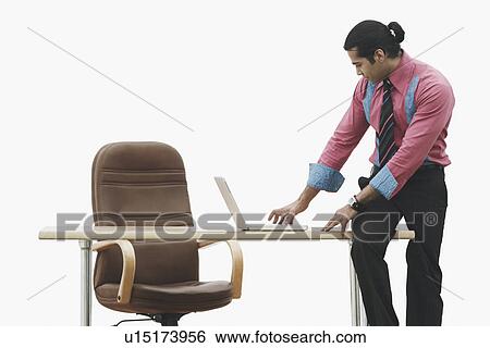 Stock Photograph - Side profile of a businessman sitting on a table working on a laptop. Fotosearch