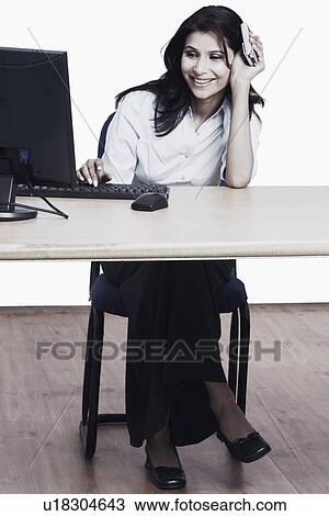 Businesswoman sitting in front of a computer holding a mobile phone View Large Photo Image Stock Image - Businesswoman sitting in front of a computer holding a mobile phone. Fotosearch
