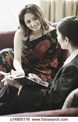 Stock Photograph - Two young women sitting on a couch discussing. Fotosearch