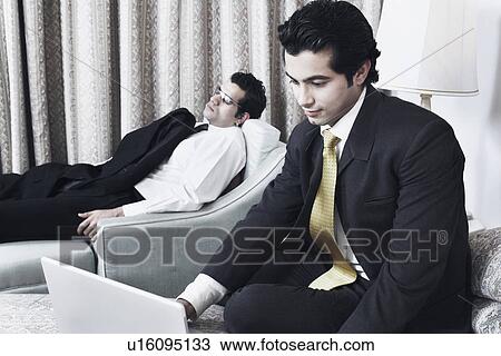 Stock Image - Close-up of a businessman using a laptop with another businessman sleeping behind him. Fotosearch