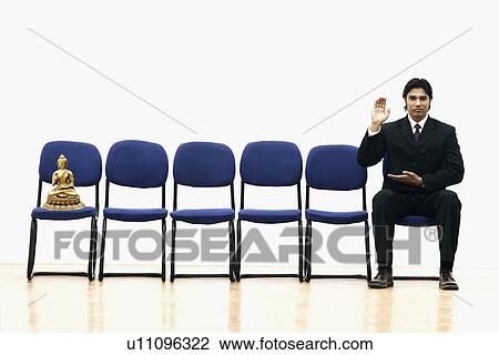Stock Image - Portrait of a businessman sitting on a chair with a statue of Buddha on a chair beside him. Fotosearch