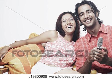 Young man holding a mobile phone with a young woman sitting on a couch View Large Photo Image Stock Photography - Young man holding a mobile phone with a young woman sitting on a couch. Fotosearch