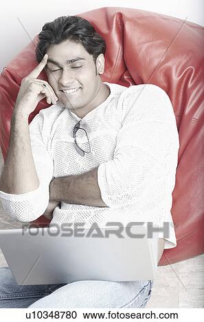Young man sitting on a bean bag in front of a laptop View Large Photo Image Stock Image - Young man sitting on a bean bag in front of a laptop. Fotosearch