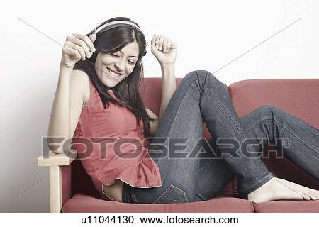 Stock Image - Young woman sitting on a couch wearing headphones and listening to music. Fotosearch
