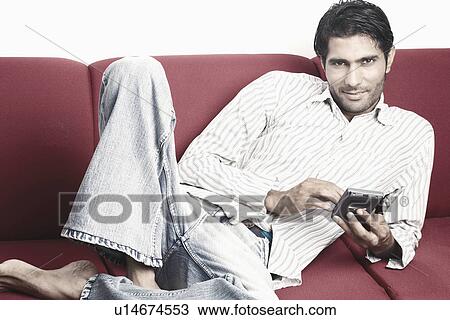 Stock Image - Portrait of a young man lying on a couch holding a personal data assistant. Fotosearch