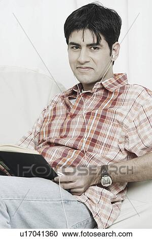 Stock Image - Portrait of a young man with a book on his lap listening to an MP3 player. Fotosearch