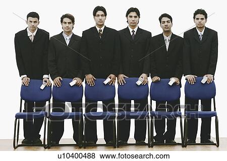 Stock Photo - Portrait of a group of young men standing behind chairs holding mobile phones. Fotosearch
