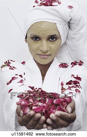 Stock Image - Portrait of a young woman with a facial mask holding rose petals. Fotosearch