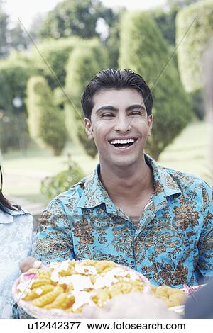Stock Photo - Portrait of a young man holding a dish of Indian sweets smiling. Fotosearch