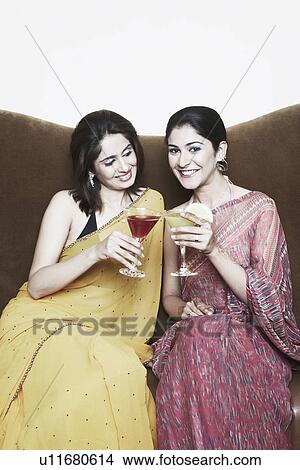 Picture - Two young women sitting on a couch toasting with martini glasses. Fotosearch