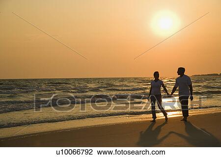 Stock Image - Caucasian mid-adult couple holding hands and walking down beach at sunset.. Fotosearch