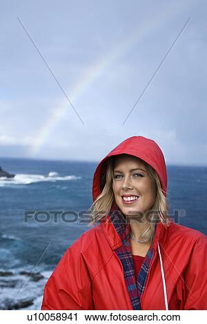 Stock Image - Caucasian mid-adult woman in red raincoat in front of ocean with rainbow in background looking at viewer and smiling in Maui, Hawaii.. Fotosearch