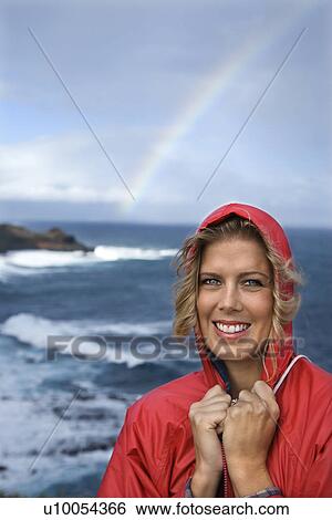 Stock Photograph - Caucasian mid-adult woman in red raincoat standing by ocean with rainbow in background in Maui, Hawaii.. Fotosearch