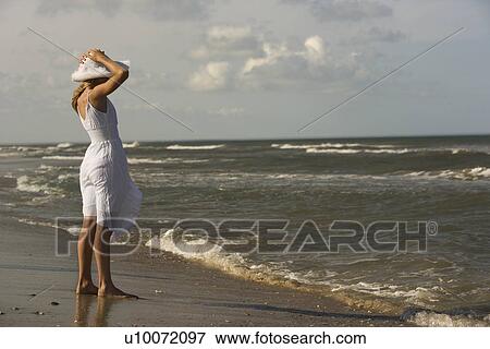 Stock Photo - Caucasian pre-teen girl standing on beach holding hat on head.. Fotosearch