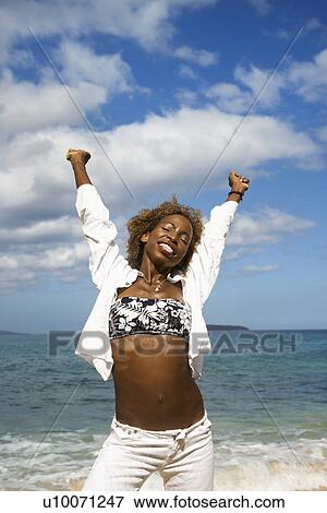 Stock Photo - Mid-adult African-American female stretching arms with ocean in background.. Fotosearch