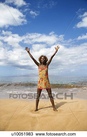 Stock Image - Mid-adult Black female with arms stretched standing in ocean.. Fotosearch