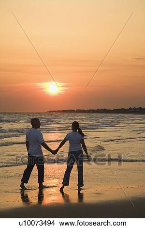 Mid-adult couple holding hands and walking on beach at sunset. View Large Photo Image Picture - Mid-adult couple holding hands and walking on beach at sunset.. Fotosearch
