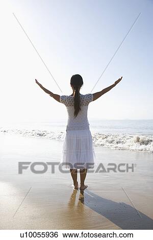 Stock Photograph - Young Asian female holding arms up standing at the shoreline.. Fotosearch