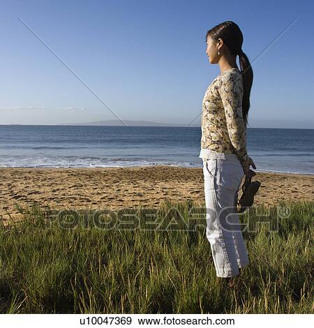 Stock Photo - Young Asian female walking on beach and carrying her sandals.. Fotosearch