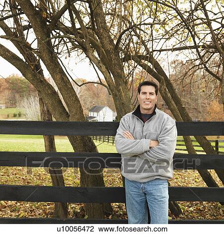 Stock Image - Caucasian man leaning against fence smiling in rural setting.. Fotosearch