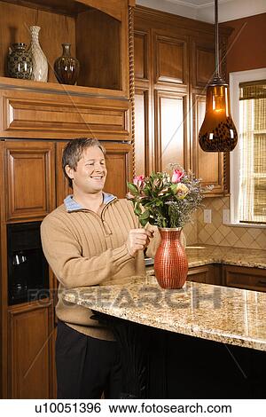 Stock Photograph - Caucasian man smiling and arranging flowers in vase in kitchen.. Fotosearch