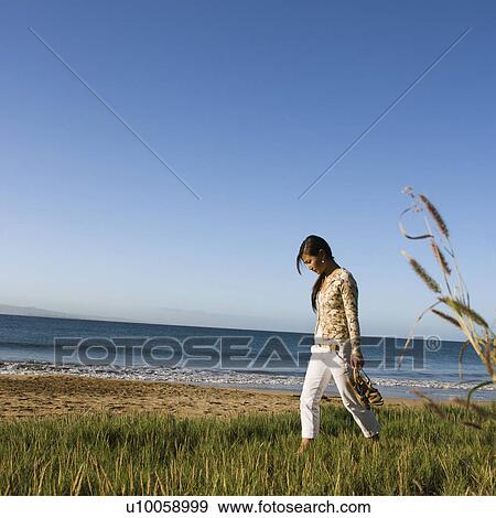 Stock Photo - Young Asian female walking on beach and carrying her sandals.. Fotosearch