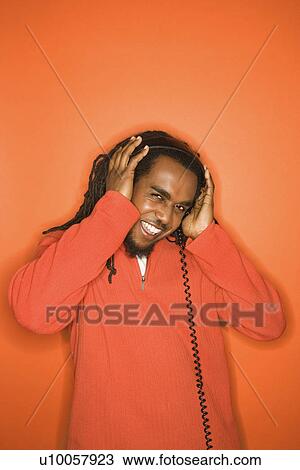 Stock Image - African-American mid-adult man listening to headphones wearing orange clothing on orange background.. Fotosearch