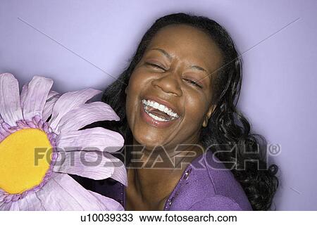 Stock Image - Portrait of African-American mid-adult woman smiling big with eyes closed holding big purple fake flower on purple background.. Fotosearch