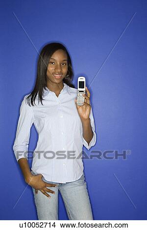 Picture - Portrait of African-American teen girl holding cellphone and hand in pocket standing against blue background.. Fotosearch