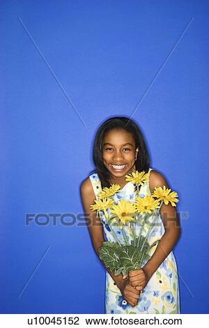 Stock Image - Portrait of African-American teen girl holding a bouquet of daisies standing in front of blue background.. Fotosearch
