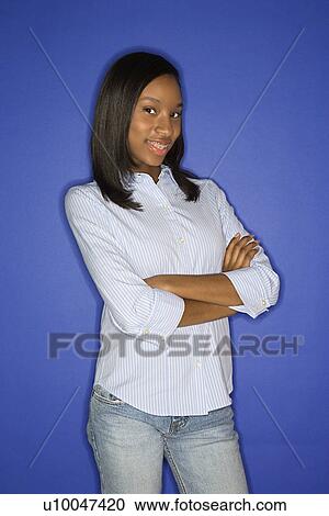 Stock Image - Portrait of smiling African-American teen girl with crossed arms standing in front of blue background.. Fotosearch