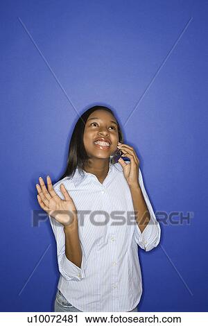Stock Image - Portrait of smiling African-American teen girl looking up and talking on cellphone standing against blue background.. Fotosearch