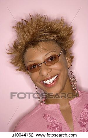 Stock Image - Portrait of smiling young African-American adult woman on pink background wearing sunglasses.. Fotosearch